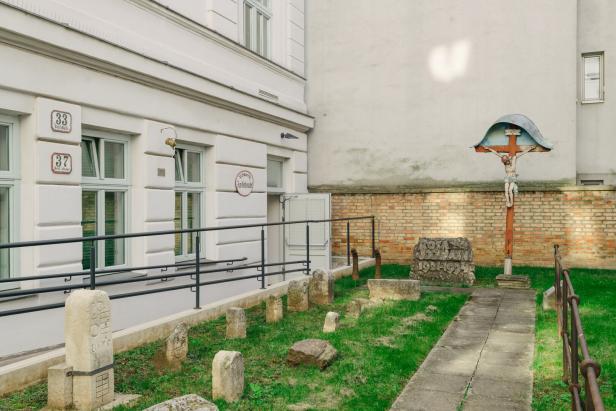 Mehrere alte Grabsteine und ein Kruzifix mit Jesusfigur stehen auf einer kleinen, umzäunten Grünfläche neben einem Gebäude.