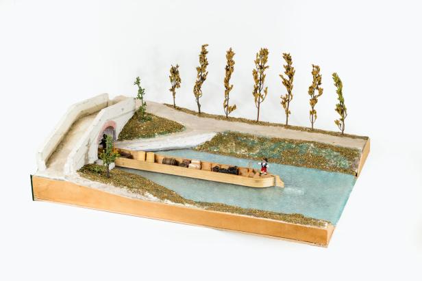 Ein Modell zeigt einen Kanal mit einem beladenen Lastkahn, einer Brücke und mehreren Bäumen am Ufer.