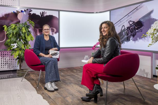 Shlomit Butbul Zwei Frauen sitzen sich in einem modernen Studio auf roten Sesseln gegenüber und unterhalten sich freundlich.
