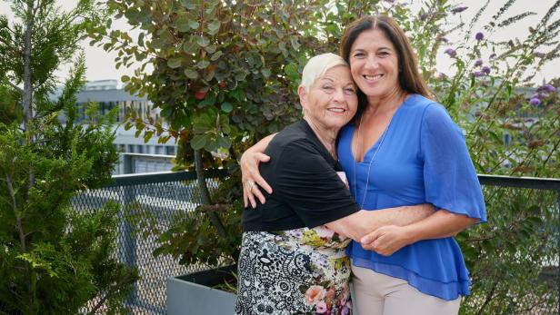 Jazz Gitti und Shlomit Butbul Zwei Frauen umarmen sich lächelnd im Freien vor grünen Pflanzen.