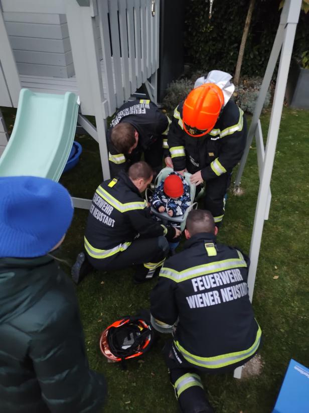 Mehrere Feuerwehrmänner stehen neben einem Schaukelgerüst inklusive Rutsche, in ihrer Mitte befindet sich ein in einer Plastikschaukel eingeklemmter Bub.