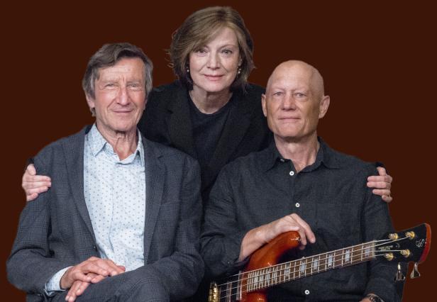 Drei Personen posieren vor einem braunen Hintergrund, ein Mann hält eine Bassgitarre.