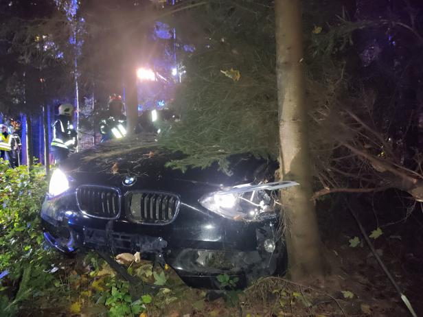 Ein schwarzes Auto ist frontal gegen einen Baum gefahren, Feuerwehrleute stehen im Hintergrund.