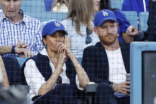 Meghan und Harry, beide mit LA-Kappen, sitzen bei einem Baseballspiel hinter einem Netz.