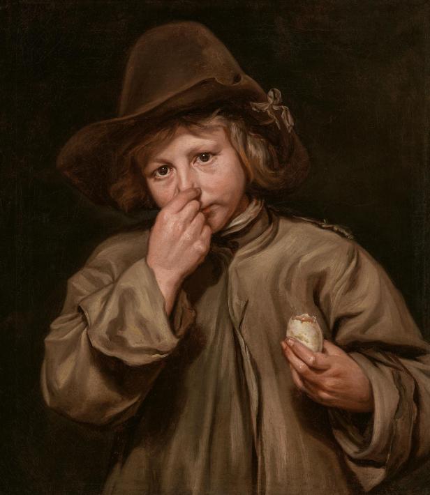 Portrait eines Jungen mit braunem, breitkrempigem Hut und locker sitzendem braunem Mantel hält ein Ei in der Hand und hält sich die Nase zu.