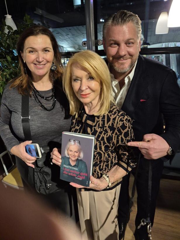 Drei Personen lächeln, eine Frau in der Mitte hält ein Buch mit dem Titel „Und weiter geht die wilde Jagd“.