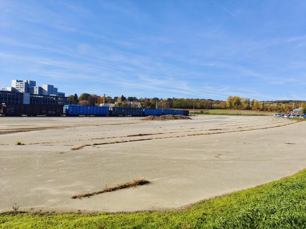 Großer leerer Platz mit vereinzeltem Gras, im Hintergrund blaue Container, Gebäude und herbstliche Bäume unter blauem Himmel.