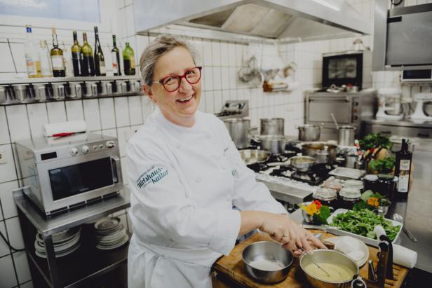 Eine Köchin mit Brille und weißer Kochjacke steht lächelnd in einer professionellen Küche und bereitet Speisen zu.