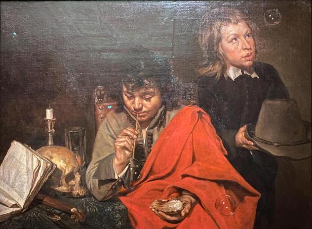 Zwei Buben in historischer Kleidung, einer hält eine Muschel und macht Seifenblasen, der andere hält einen Hut in seinen Händen. Totenkopf, Kerze und Buch sind auf dem Tisch daneben.