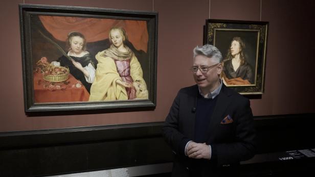 Mann in dunklem Anzug vor zwei klassischen Porträtgemälden an dunkelroter Wand.