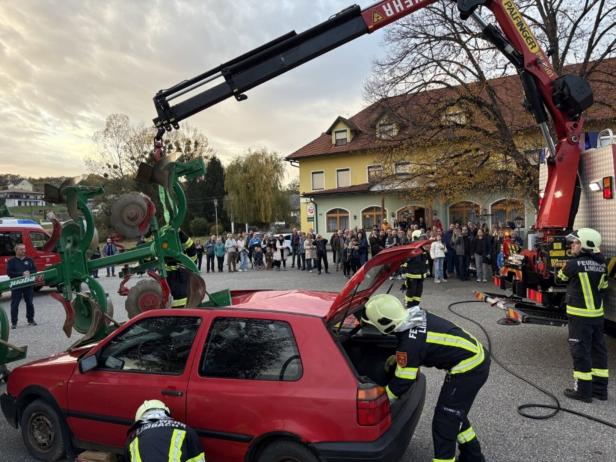 Feuerwehrleute bergen mit einem Kran ein landwirtschaftliches Gerät von einem roten Auto, während viele Menschen zuschauen.