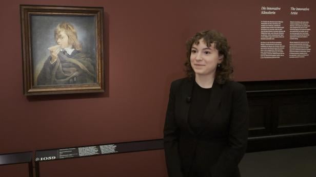 Person mit lockigem Haar und schwarzer Kleidung steht vor einer Wand mit einem gerahmten Gemälde und Texttafeln.