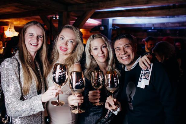 Vier Personen stehen eng zusammen, lächeln und stoßen mit goldenen Sektgläsern an.