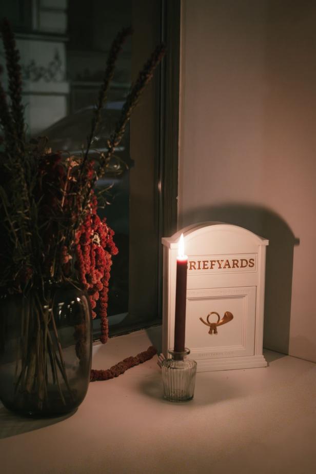 Griefyards Ausstellung Kerze, Trockenblumen und ein weißer Briefkasten mit der Aufschrift "GRIEFYARDS" auf einer Fensterbank.