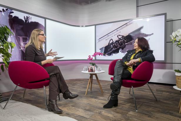 Sabine Holzinger Zwei Frauen sitzen in roten Sesseln und unterhalten sich in einem modernen Studio mit großen Bildschirmen im Hintergrund.