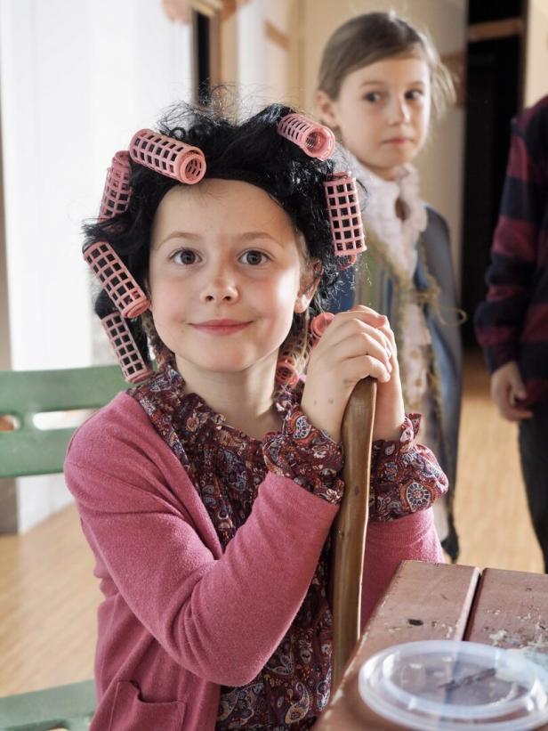 Ein Kind mit Lockenwicklern im Haar und bunter Kleidung sitzt lächelnd an einem Tisch, im Hintergrund steht ein weiteres Kind.