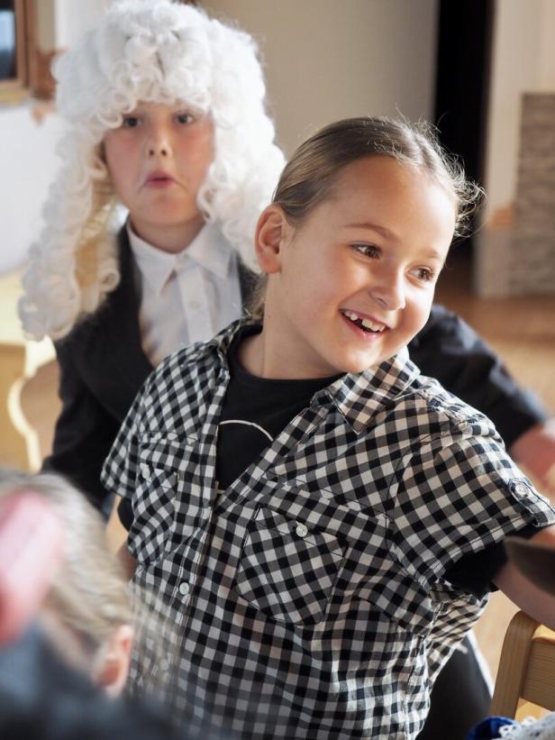 Zwei Kinder spielen, eines trägt eine weiße Perücke und Anzug, das andere lacht und trägt ein kariertes Hemd.