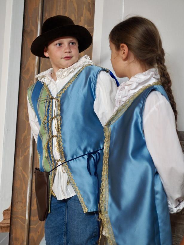 Zwei Kinder in historischen Kostümen mit blauen Westen und weißen Hemden stehen nebeneinander und schauen sich an.