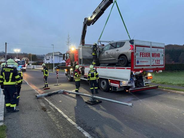 Mehrere Feuerwehrleute bergen ein beschädigtes Auto mit einem Kran, während ein umgestürzter Laternenmast auf der Straße liegt.