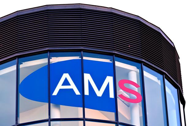 Großes AMS-Logo auf einer Glasfassade mit blauer und roter Schrift, darüber schwarze Lamellen.