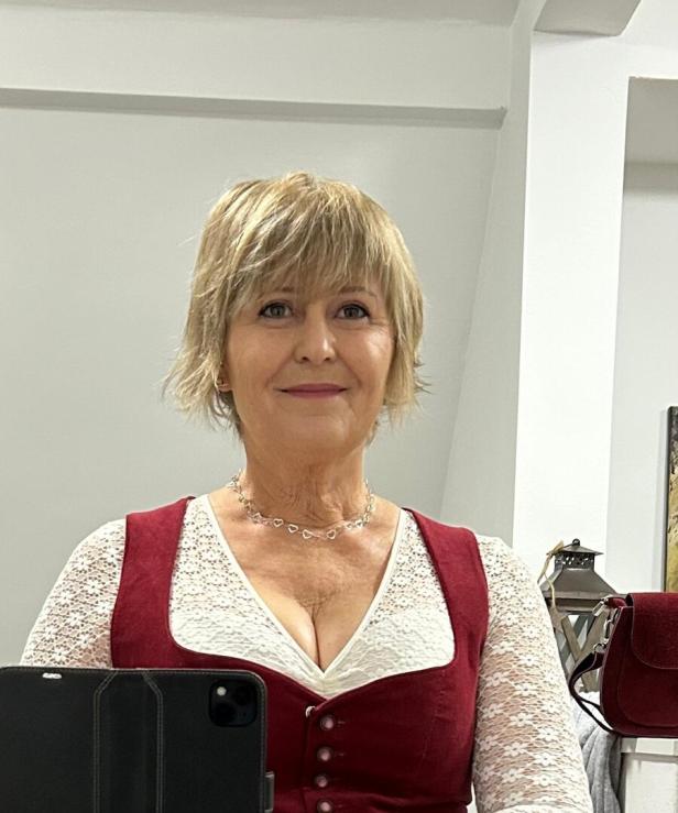 Eine Frau mit kurzem, blondem Haar trägt ein rotes Dirndl mit weißer Spitzenbluse und lächelt in die Kamera.