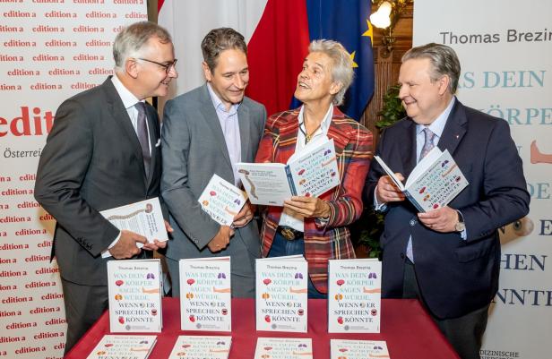 Vier Personen im Anzug stehen lachend zusammen und halten ein Buch mit dem Titel „Was dein Körper sagen würde, wenn er sprechen könnte“.