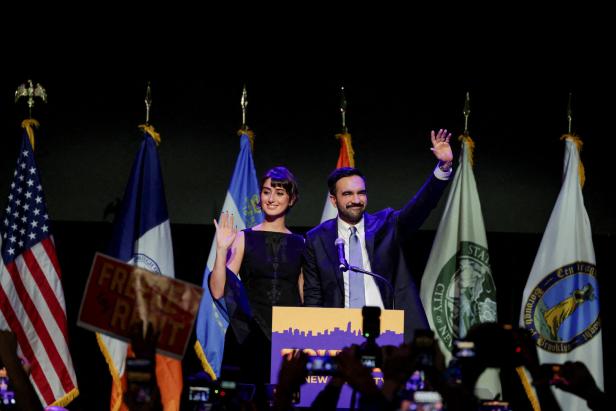 Ein Mann und eine Frau stehen lächelnd auf einer Bühne, winken dem Publikum zu und sind von mehreren Flaggen umgeben.