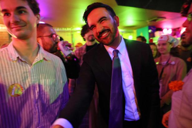 Ein Mann im Anzug lächelt breit und streckt die Hand aus, umgeben von fröhlichen Menschen in bunter Beleuchtung.