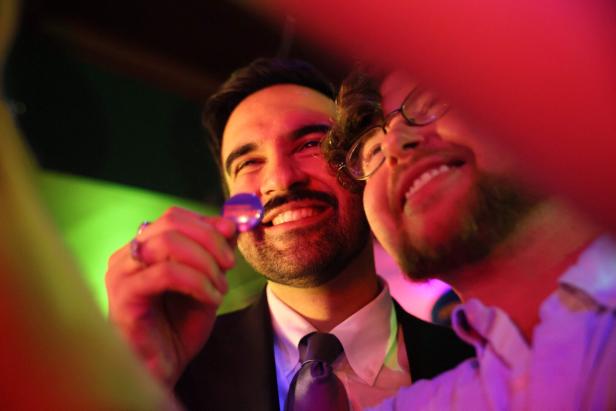 Zwei lachende Männer stehen eng beieinander in bunter Beleuchtung, einer hält einen kleinen Gegenstand in der Hand.