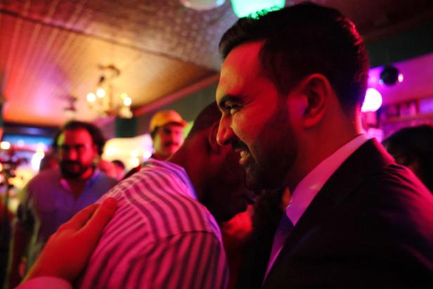 Ein Mann im Anzug umarmt herzlich eine andere Person in einer bunt beleuchteten, belebten Bar.