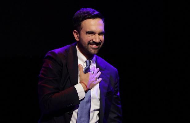 Ein Mann im Anzug lächelt und legt eine Hand dankbar auf seine Brust, während er auf einer dunklen Bühne steht.