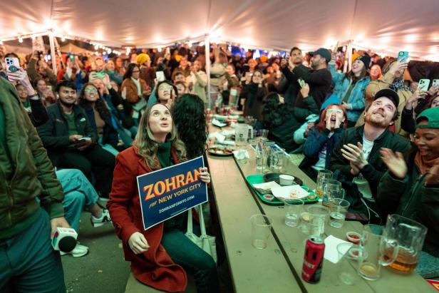 Viele Menschen sitzen und stehen unter einem Zelt, jubeln und halten Schilder und Handys, eine Frau hält ein Wahlkampfschild.