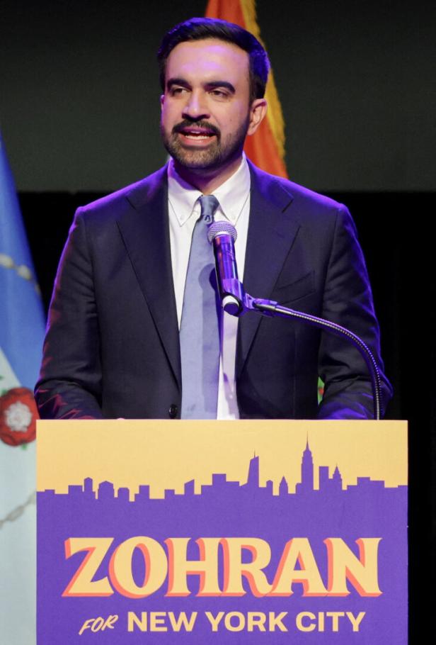 Ein Mann im Anzug spricht an einem Rednerpult mit der Aufschrift „Zohran for New York City“ in lila und gelb.