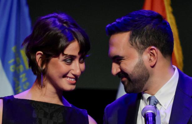 Zwei Personen stehen nebeneinander, lächeln sich an und wirken glücklich, im Hintergrund sind Fahnen zu sehen.