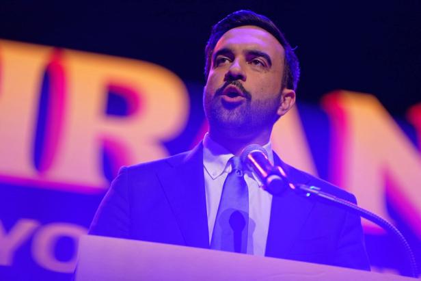 Ein Mann im Anzug spricht an einem Mikrofon vor einem farbig beleuchteten Hintergrund.