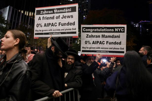 Mehrere Menschen stehen nachts zusammen, einige halten große Schilder mit politischer Botschaft und loben Zohran Mamdani.