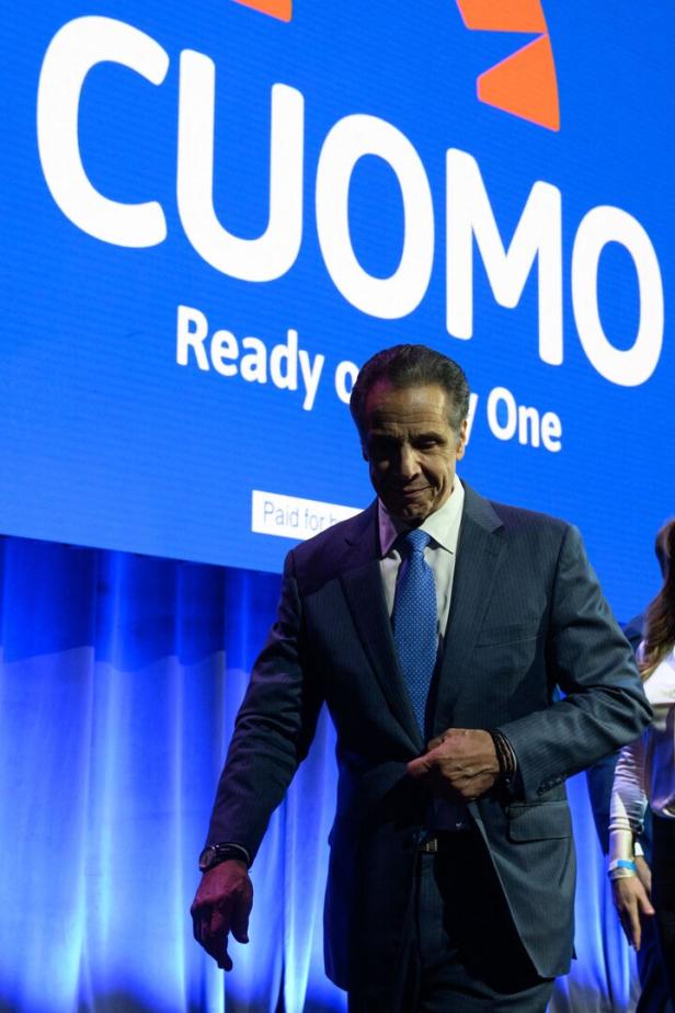 Ein Mann im Anzug steht vor einer großen blauen Leinwand mit dem Schriftzug „CUOMO Ready on Day One“.