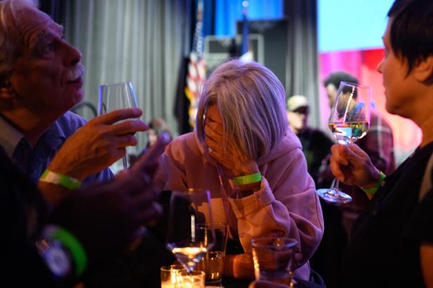 Eine Frau sitzt mit gesenktem Kopf und verdecktem Gesicht zwischen Menschen, die Weingläser halten, und wirkt emotional betroffen.