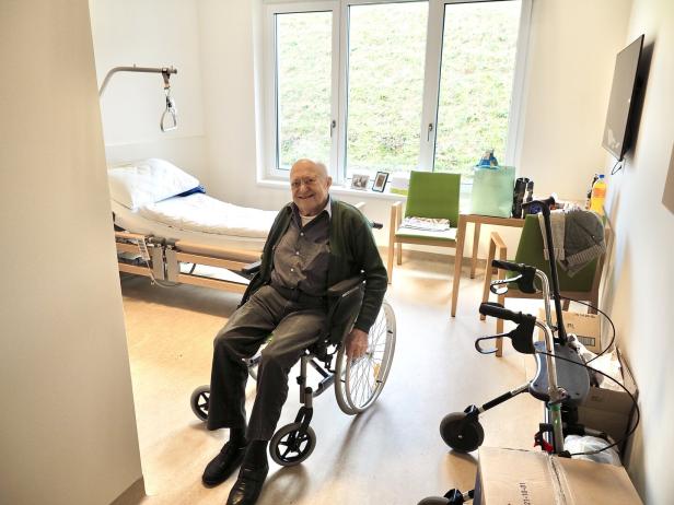 Ein älterer Mann sitzt lächelnd im Rollstuhl in einem hellen, aufgeräumten Pflegezimmer mit Pflegebett und Gehhilfe.