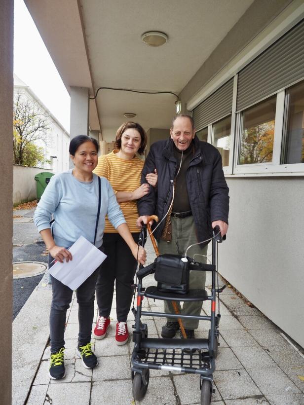 Drei Personen stehen draußen, eine Frau hält einen Zettel, ein Mann mit Rollator wird von einer weiteren Frau gestützt.
