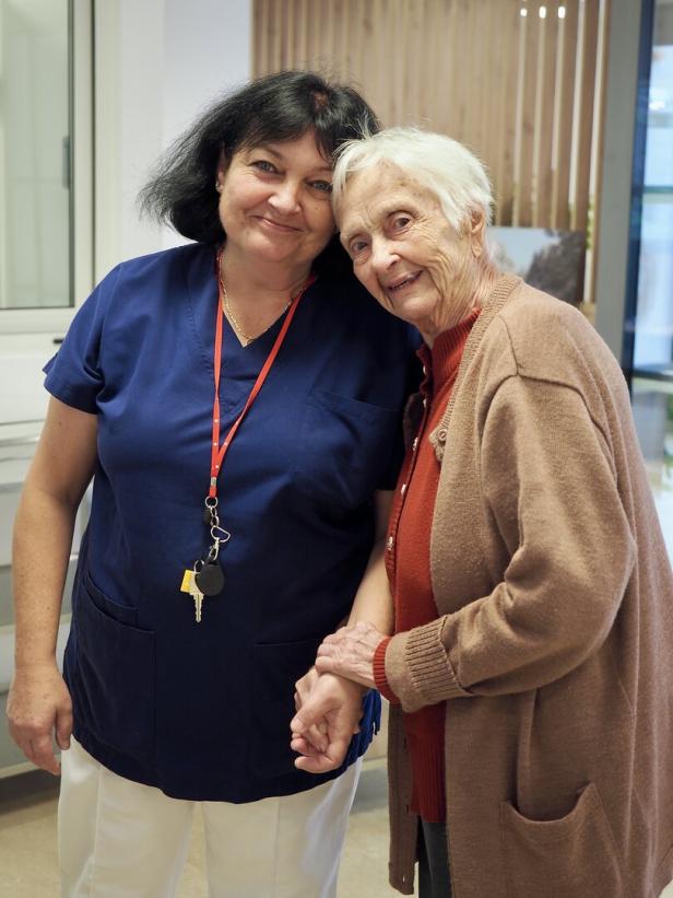 Eine ältere Frau und eine Pflegekraft stehen lächelnd nebeneinander und halten sich an den Händen.