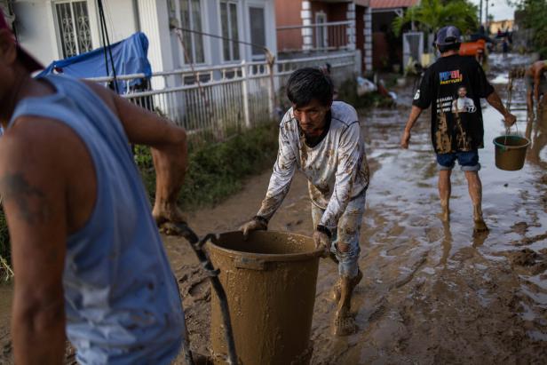Mehrere Menschen räumen eine schlammige Straße auf, indem sie Eimer und Mülltonnen voller Schlamm transportieren.