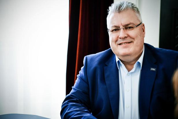 Michael Mertin lächelt in die Kamera. Er trägt ein hellblaues Hemd und einen dunkelblauen Anzug. Hinter ihm sieht man einen dunkelroten Vorhang.
