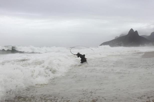 Person in dunkler Kleidung steht im Wasser am Strand und schwingt einen langen, gebogenen Gegenstand, im Hintergrund bewölkter Himmel und Berge.