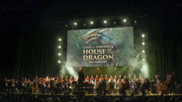 Orchester auf Bühne mit Dirigent vor großer Leinwand, die einen Drachen und den Text 'GAME OF THRONES & HOUSE OF THE DRAGON DAS KONZERT' zeigt.