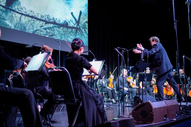 Dirigent vor einem Orchester mit Streichinstrumenten auf der Bühne, im Hintergrund eine Projektion mit Stacheldraht und Himmel.