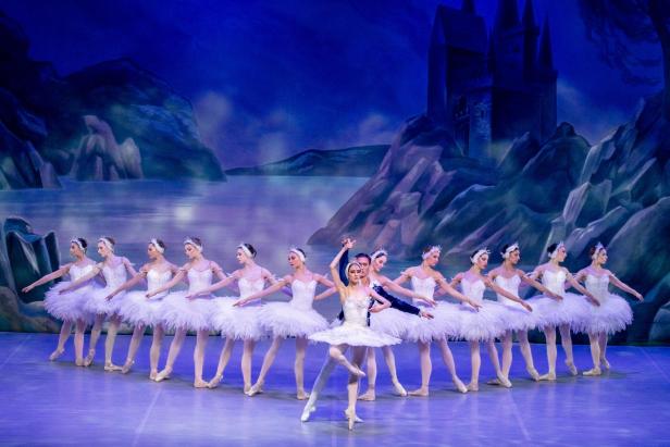 Balletttänzerinnen in weißen Tutus stehen in einer Reihe, zwei Tänzer in der Mitte führen eine Pose auf einer Bühne mit Schlosskulisse aus.