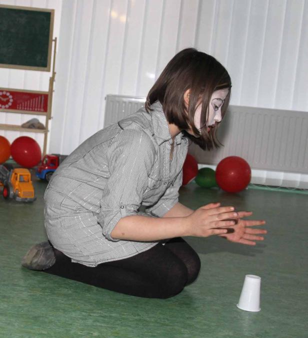 Ein Mädchen mit weißer Gesichtsbemalung kniet vor einem Plastikbecher.