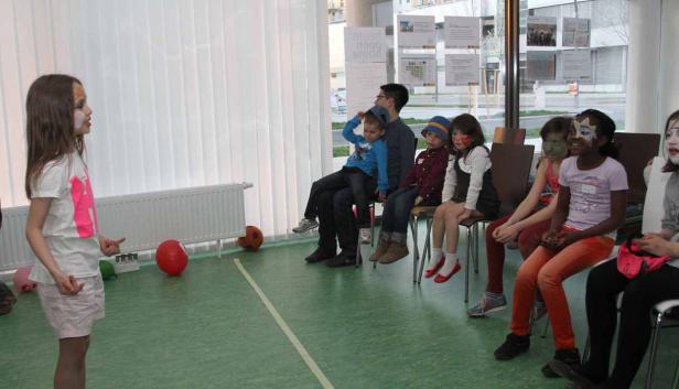 Eine Gruppe von Kindern mit Gesichtsbemalung sitzt und schaut einer sprechenden jungen Frau zu.