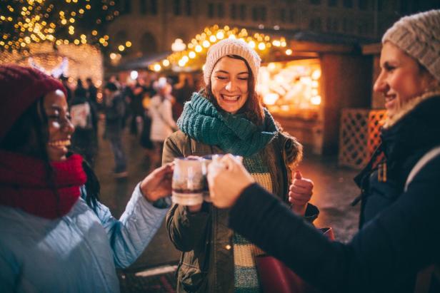 Drei Frauen auf einem Weihnachtsmarkt prosten sich mit einem Winterbecher  zu und lächeln sich an.